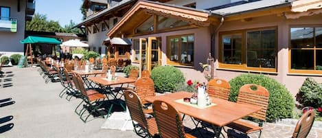 Un'area pranzo all'aperto con tavoli e sedie in legno, una zona salotto coperta e un edificio con ampie finestre e balconi.