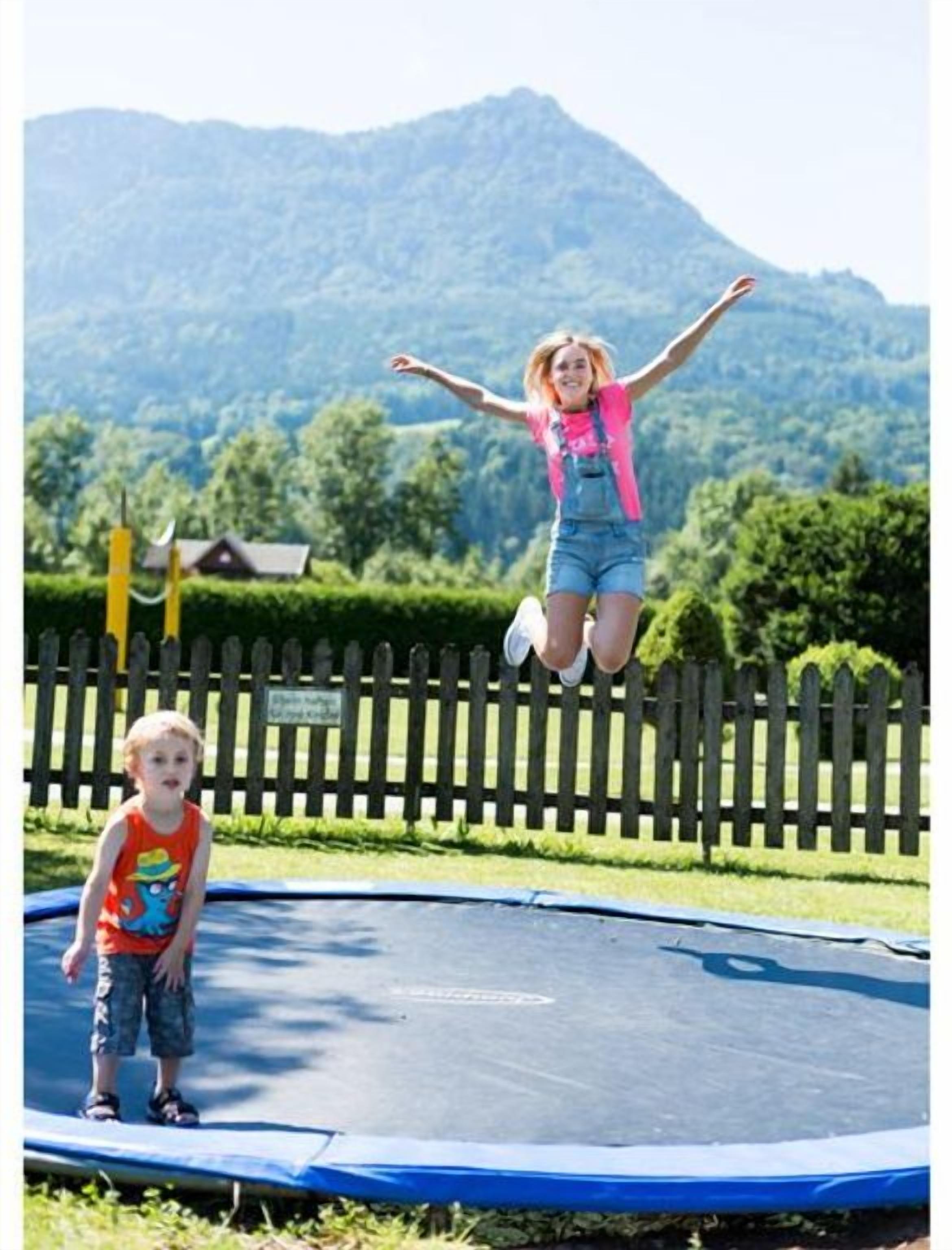 Un trampolín con un niño saltando y otro de pie, rodeado de una valla de madera y follaje.