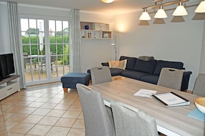 A modern living room with a sofa, a dining table, and chairs. There is a television on the left and a bookshelf with various items.