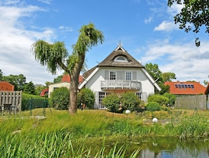 A white house with a balcony, surrounded by greenery and a pond.