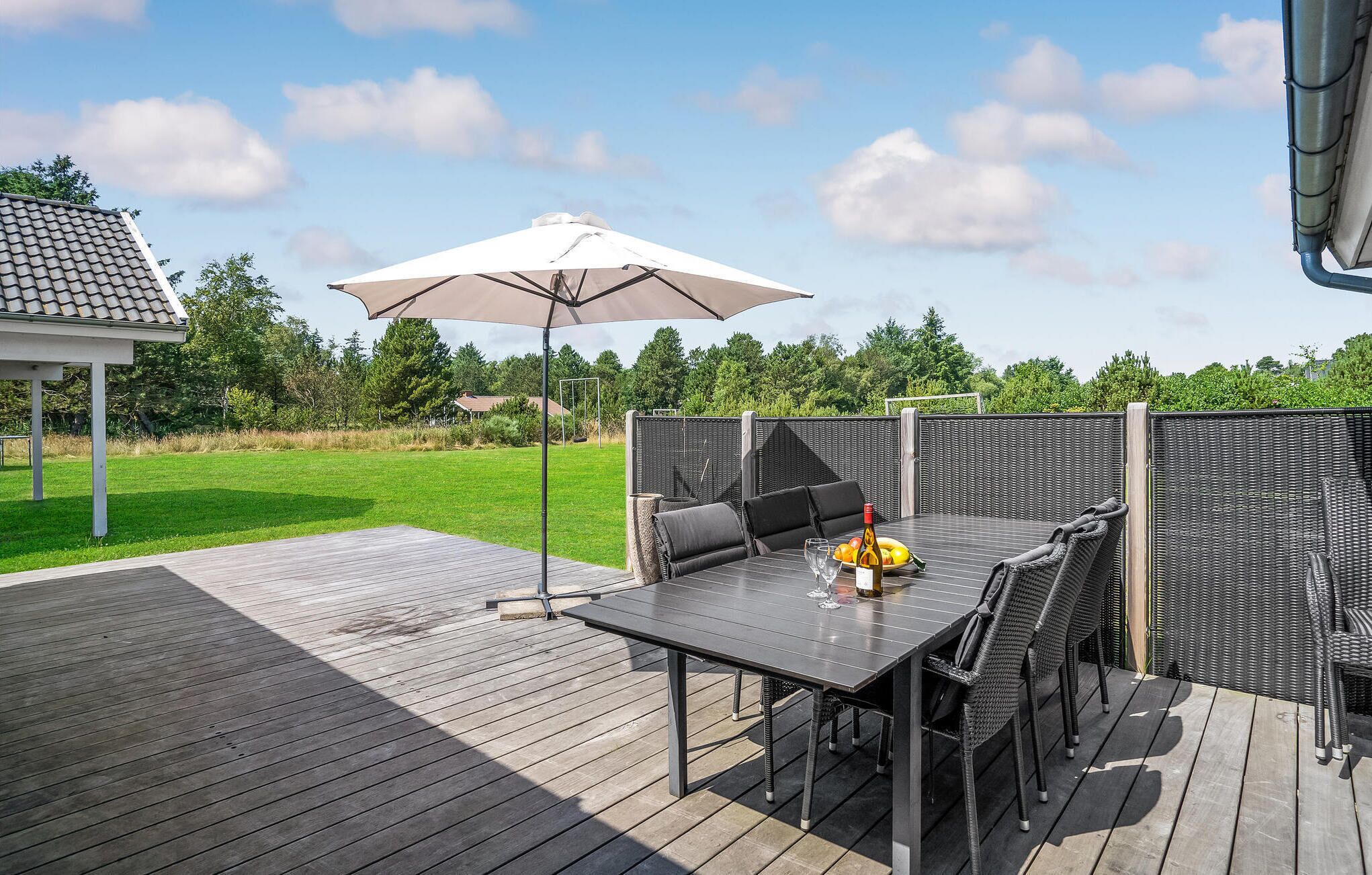 En udendørs terrasse med et bord og stole, en pergola med parasol samt udsigt til en græsplæne og træer.