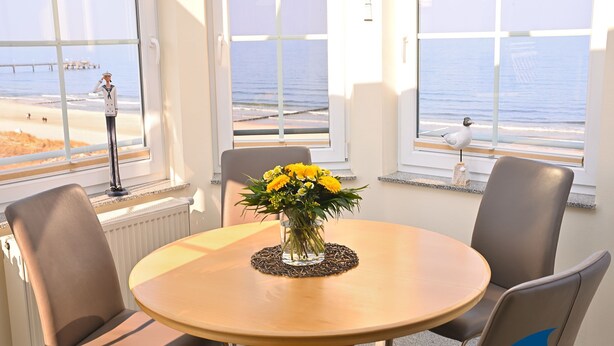 Et rundt bord med en vase med gule blomster, fire stole og havudsigt gennem store vinduer.