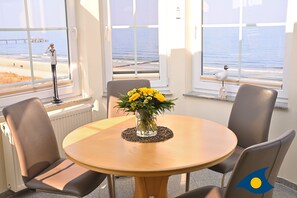 Uma mesa redonda com um vaso de flores amarelas, quatro cadeiras e vista para a praia através de grandes janelas.