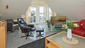 En moderne stue i loftstil med højt til loftet, et stort vindue, en rød sofa, en sort læderlænestol, et sofabord i glas og et fjernsyn.
