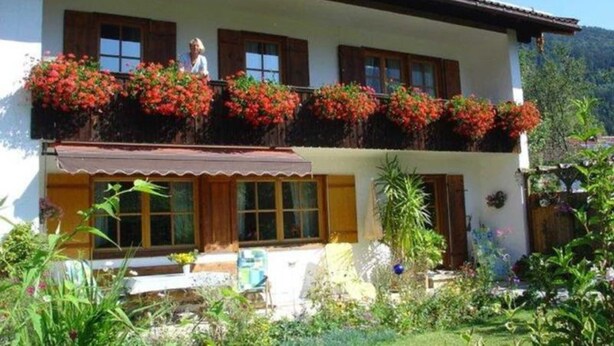Ein zweistöckiges Gebäude mit einem Balkon, der mit roten Blumen geschmückt ist, einer Veranda und einem Garten mit verschiedenen Pflanzen.