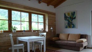 Ein gemütlicher Raum mit Holzvertäfelung, einem weißen Tisch mit Stühlen, einem braunen Sofa und einem großen Fenster mit Blick auf Grünflächen.