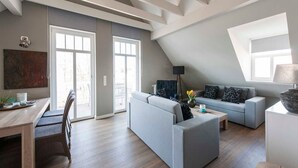 Ein geräumiges Wohnzimmer mit hoher Decke, eine Essecke mit Holztisch und -stühlen, eine graue Sofagarnitur und ein großes Fenster, das für natürliches Licht sorgt.
