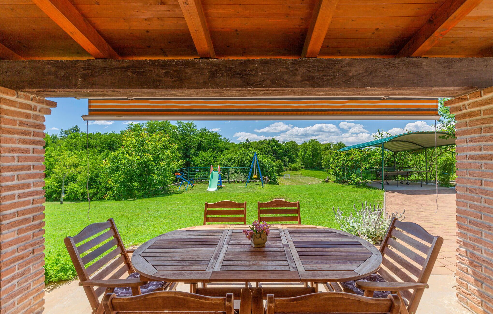 Un patio cubierto con una mesa y sillas de madera, un muro de ladrillo y vistas a un área verde con juegos infantiles.