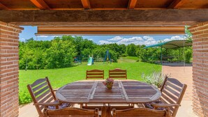 Eine überdachte Terrasse mit Holztisch und -stühlen, einer Ziegelwand und Blick auf eine Rasenfläche mit Spielgeräten.