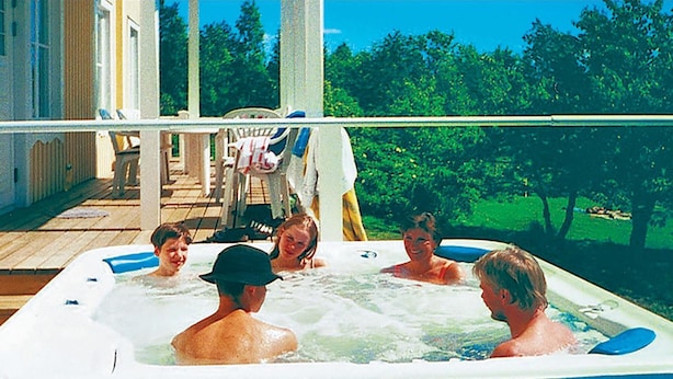 Un jacuzzi con cinco personas, rodeado por una terraza de madera y una casa al fondo.