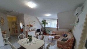 Ein Wohnzimmer mit Esstisch, Stühlen, einer Couch und einem Fernseher. Übertöpfe und eine Deckenleuchte sind ebenfalls vorhanden.