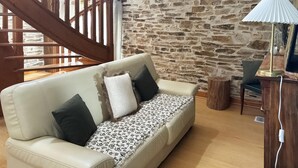 Ein gemütliches Zimmer mit Steinwand, einer Holztreppe, einem beige gefärbten Sofa mit dekorativer Tagesdecke und einer Lampe auf einem Holztisch.