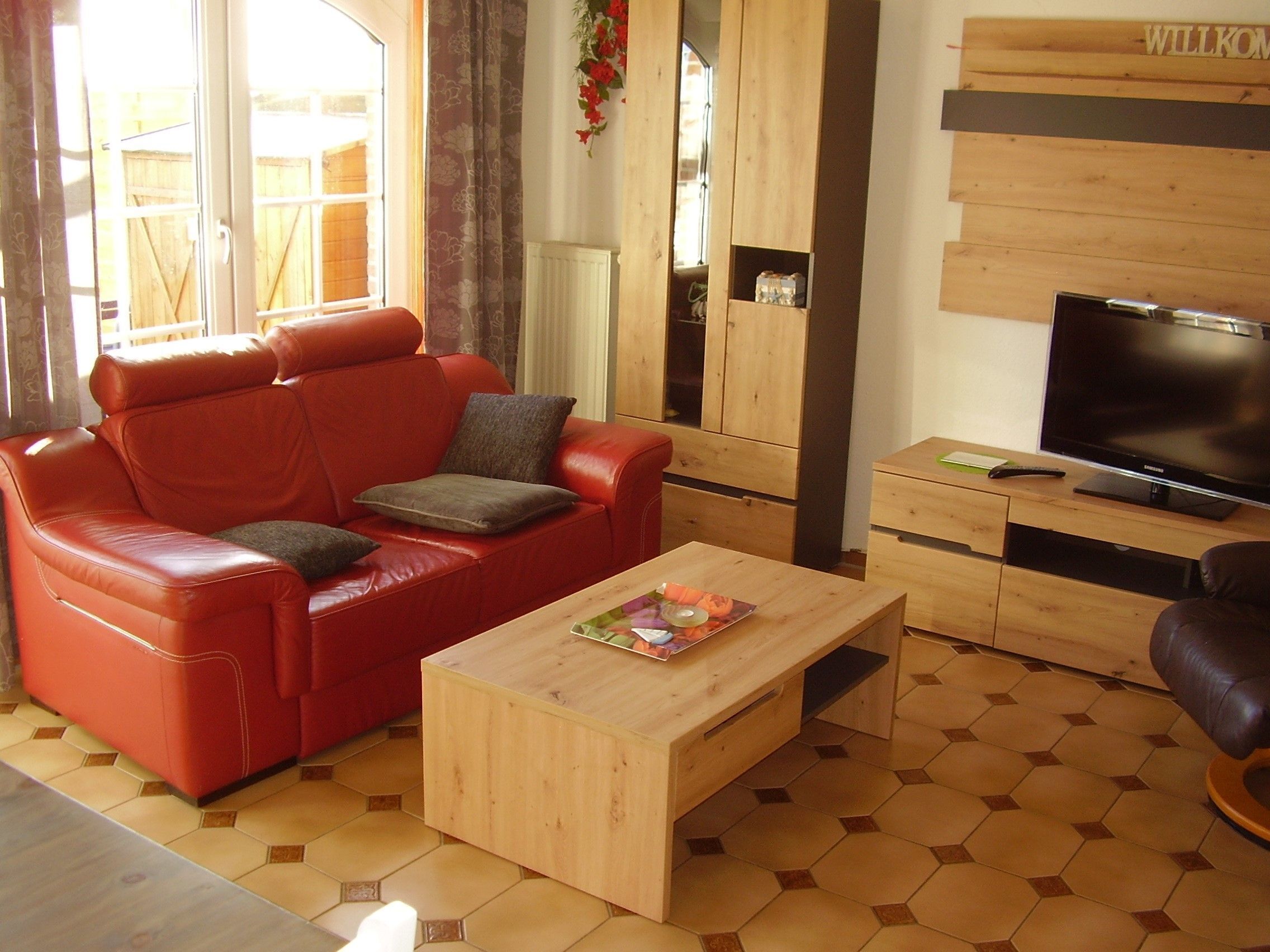 Uma sala de estar com um sofá de couro vermelho, uma mesa de centro de madeira, uma TV e um armário de madeira com uma placa 'WILKOM'.