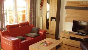 Ein Wohnzimmer mit einem roten Ledersofa, einem Couchtisch aus Holz, einem Fernseher und einer Holzkassette mit dem Schriftzug 'WILKOM'.