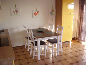 Un coin repas avec une table en bois, des chaises blanches et des carreaux de sol à motifs hexagonaux. Des tableaux représentant des motifs floraux, encadrés, ornent le mur.