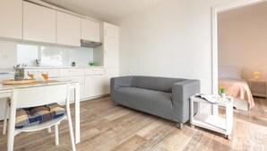 Un espace de vie moderne et minimaliste, avec un canapé gris, une petite table blanche et un coin cuisine doté de meubles blancs.