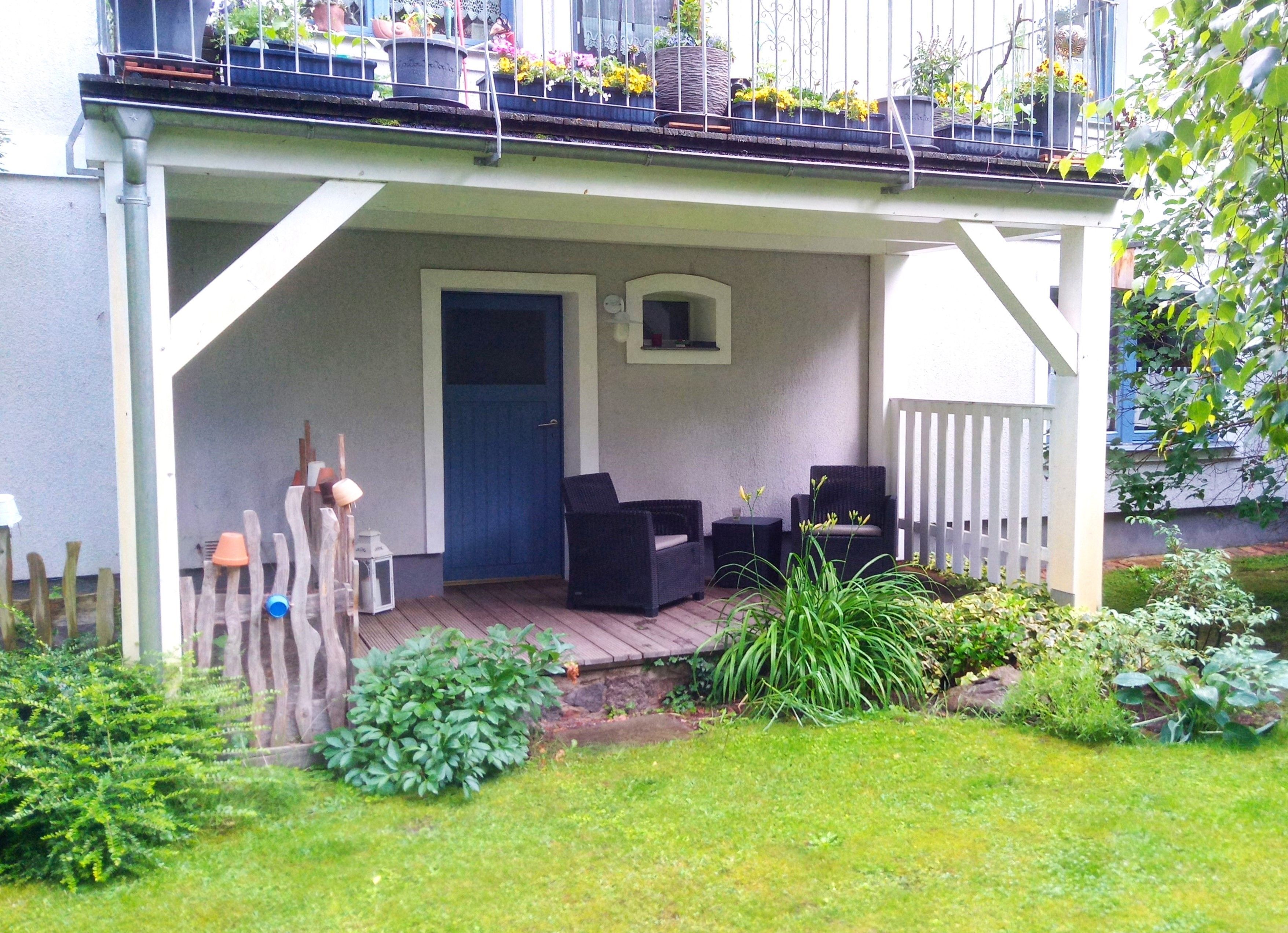 Een overdekte veranda met twee zwarte wicker stoelen en een kleine tafel, een blauwe deur en een witte hekken.
