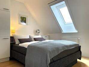 A bedroom with a bed, a bedside table with a lamp, a fan, a radiator, and a skylight.
