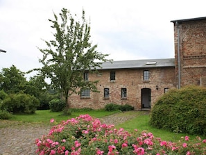 Un bâtiment en briques à l’architecture traditionnelle, un chemin bordé de pierres et un jardin agrémenté de fleurs roses.