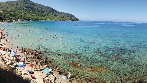 Ein Strand mit klarem Wasser, felsigem Ufer und einem Berg im Hintergrund.