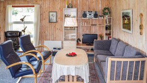 A cozy cabin interior with wooden walls, a dining area with a table and chairs, a sofa, a TV, and a shelf with various items.