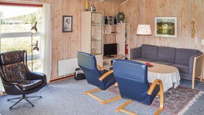 A cozy room with a wooden interior, a grey sofa, a black leather chair, and a blue armchair. There is a small table with a red flower, a bookshelf, and a framed picture on the wall.