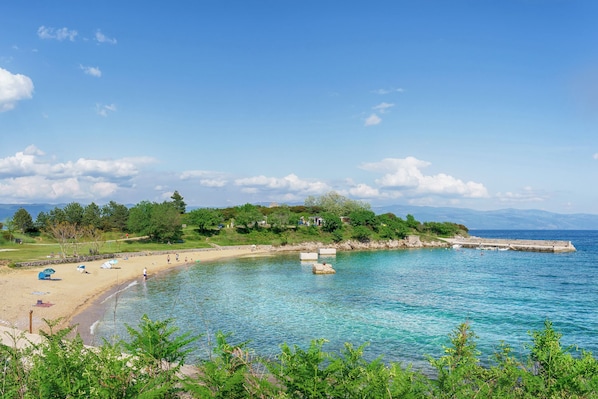 A coastal beach with clear blue water, a sandy shore, and a stone pier extending into the sea.