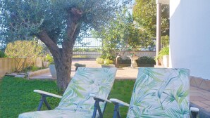Deux fauteuils de jardin ornés d’un motif de feuilles tropicales, un arbre et une terrasse dallée agrémentée de plantes en pot et d’une fontaine.