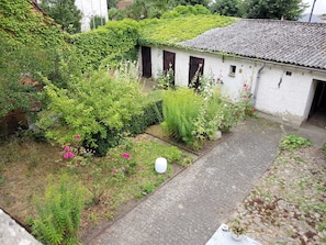 Um pátio com caminho de pedras, vegetação exuberante e um edifício branco com persianas escuras.