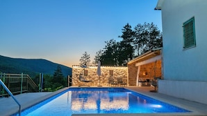Piscina con pared de piedra y zona de estar al aire libre, rodeada de árboles y una montaña al fondo.