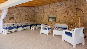 Una habitación con paredes de piedra, techo de madera, un conjunto de sofás, sillas de mimbre blanco y un televisor montado en la pared.