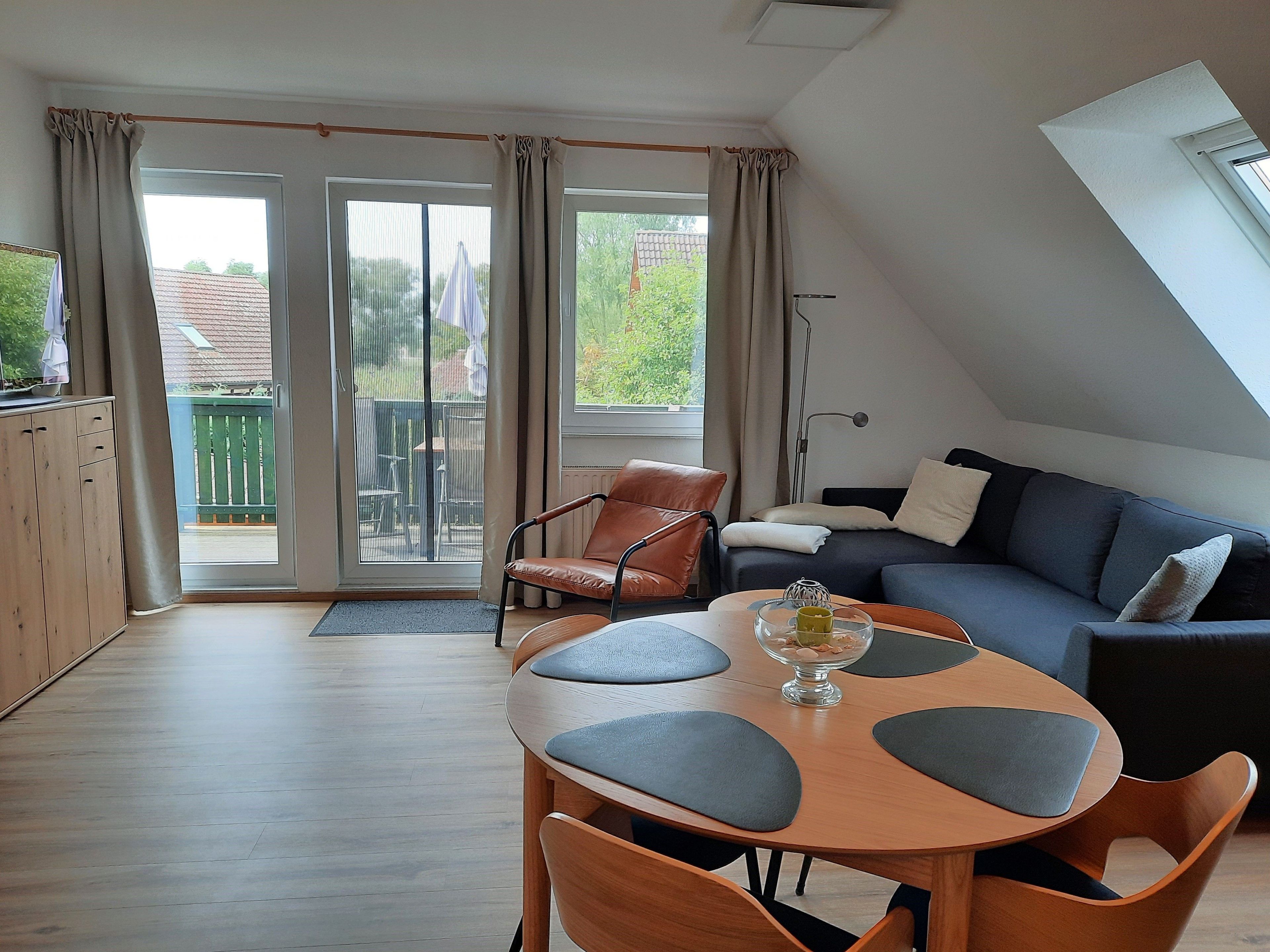 A modern living room with a round wooden table, two chairs, a sofa, and a balcony with a view of a green landscape.