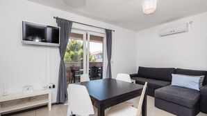 Un saló modern amb un sofà fosc, una taula de dinar amb cadires, un televisor de pantalla plana penjat a la paret i un balcó amb taula i cadires.