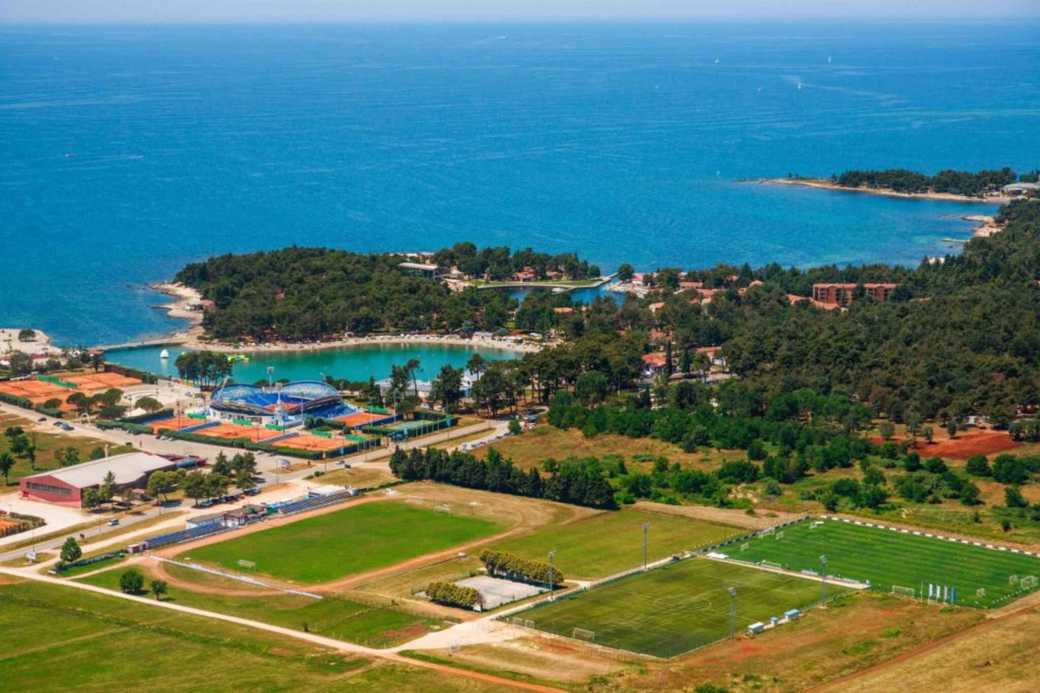 An aerial view of a coastal area with a stadium, sports fields, and a beach.