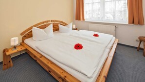 Un lit bien fait, avec du linge de lit blanc et deux fleurs rouges, une table de chevet en bois avec une lampe, une fenêtre avec des rideaux et un radiateur.