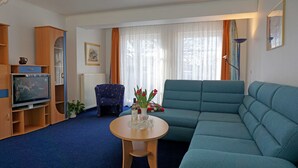 Un salon avec un canapé bleu, une petite table sur laquelle est posé un vase de fleurs, un téléviseur et une fenêtre avec des rideaux.