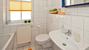 Une salle de bain moderne équipée de toilettes, d’un lavabo et d’une baignoire. On y trouve une fenêtre avec un store, un radiateur et une étagère contenant des articles de toilette.