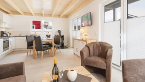 Un salon moderne avec un fauteuil en cuir marron, un coin repas avec une table et des chaises en bois, et une cuisine avec des meubles blancs et un mur d’accent rouge.