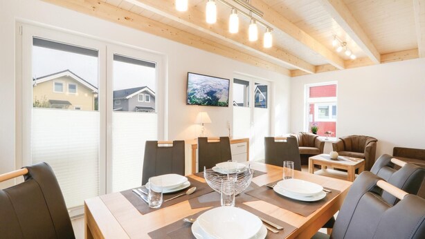 Un coin repas avec une table en bois, dressée pour quatre personnes, entourée de chaises. De grandes fenêtres se trouvent d’un côté, et un téléviseur est fixé au mur.