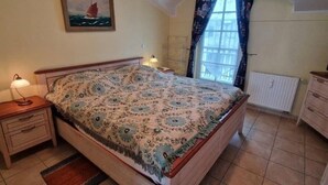 Un dormitori amb un llit, una tauleta de nit, una làmpada, una finestra amb cortines i un radiador.