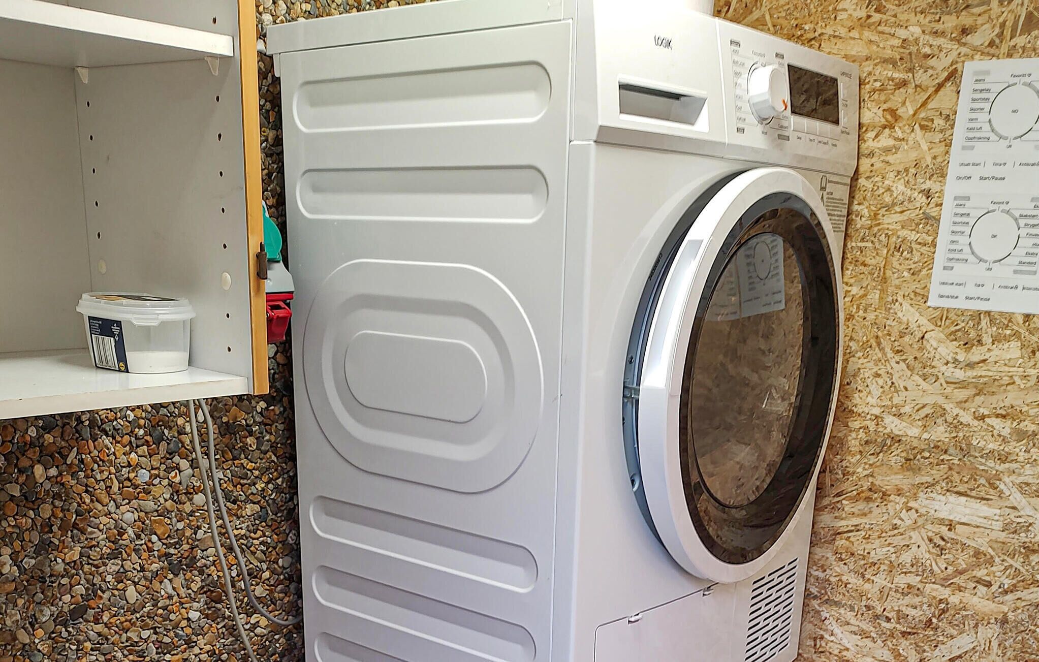 Une machine à laver à chargement frontal se trouve dans une buanderie. Au-dessus, une étagère contient un récipient à lessive et un objet rouge.