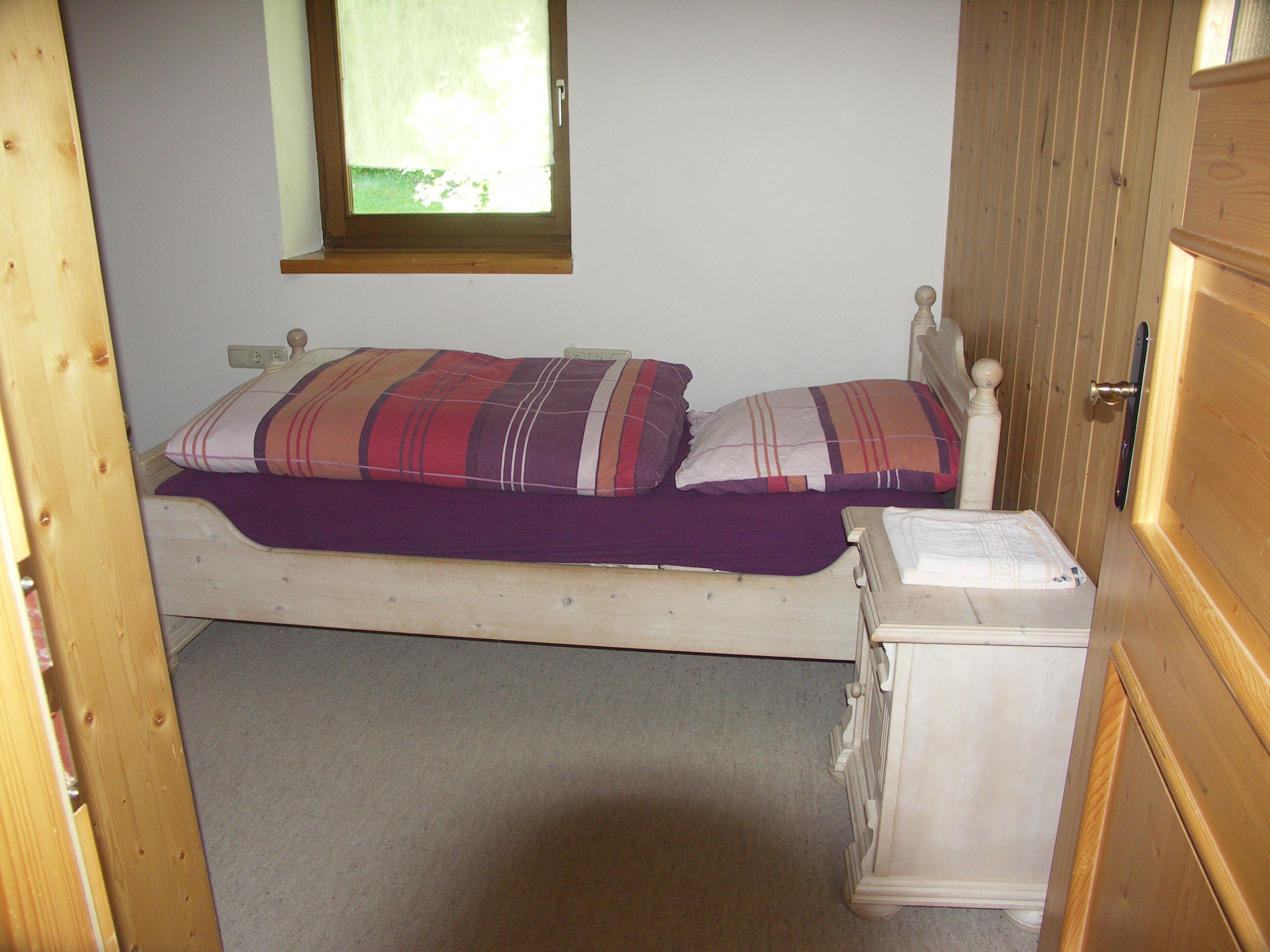 A small, single-bed room with a wooden headboard, a bedside table, and a window with a view of greenery outside.