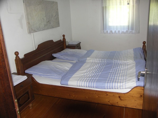 Un lit simple avec un cadre en bois, une table de chevet, une commode et une fenêtre avec des rideaux légers.