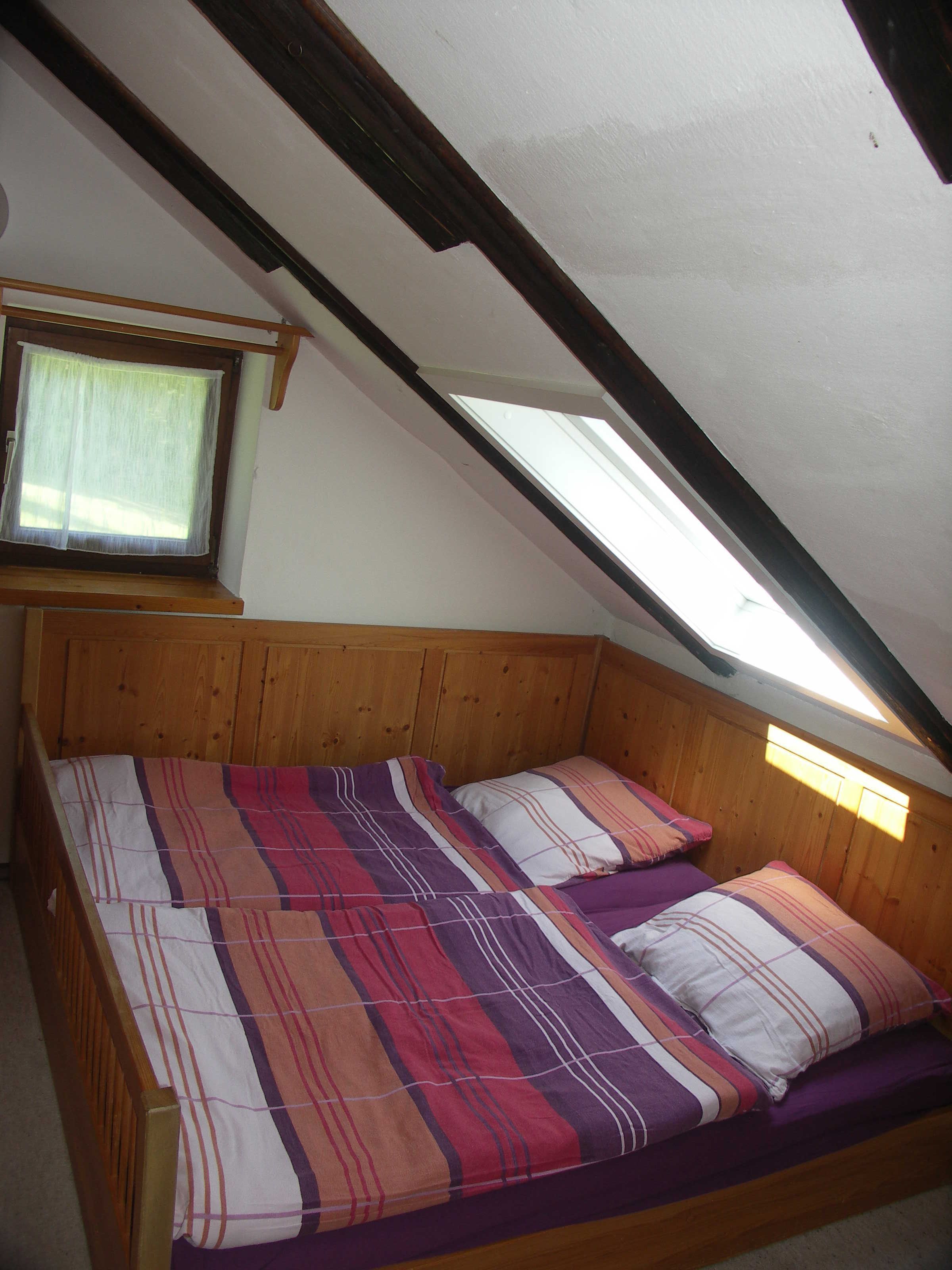A bedroom with a sloped ceiling, wooden headboard, and a window with a sheer curtain.