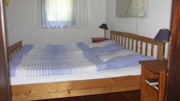 A single bed with a wooden frame, a bedside table with a lamp, a small wooden shelf, and a window with a sheer curtain.