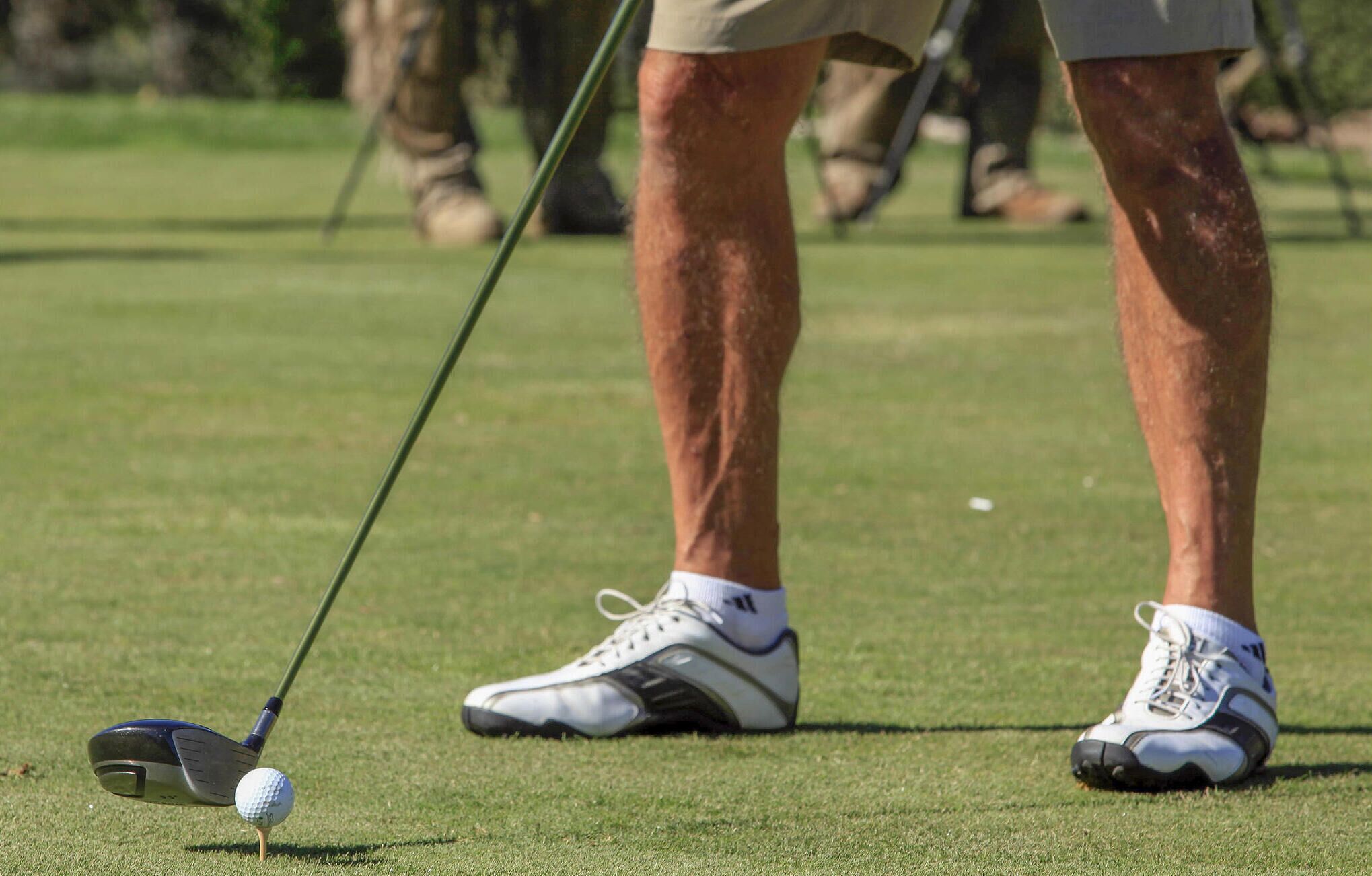 Clube de golfe posicionado sobre uma bola de golfe em um tee, com pernas e sapatos de uma pessoa à mostra.