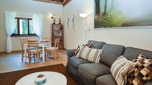 Een woonkamer met een grijze bank, een witte salontafel en een eethoek met houten stoelen en een tafel. Er staat een boekenkast met boeken en decoratieve objecten, en een schilderij van een bos aan de muur.