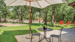 Een terras buiten met een ronde tafel en stoelen, een grote parasol en een uitzicht op een tuin met bomen en een hek op de achtergrond.