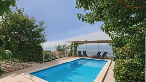 Una piscina rettangolare con bordatura in pietra, circondata da vegetazione e un pergolato con sedie.