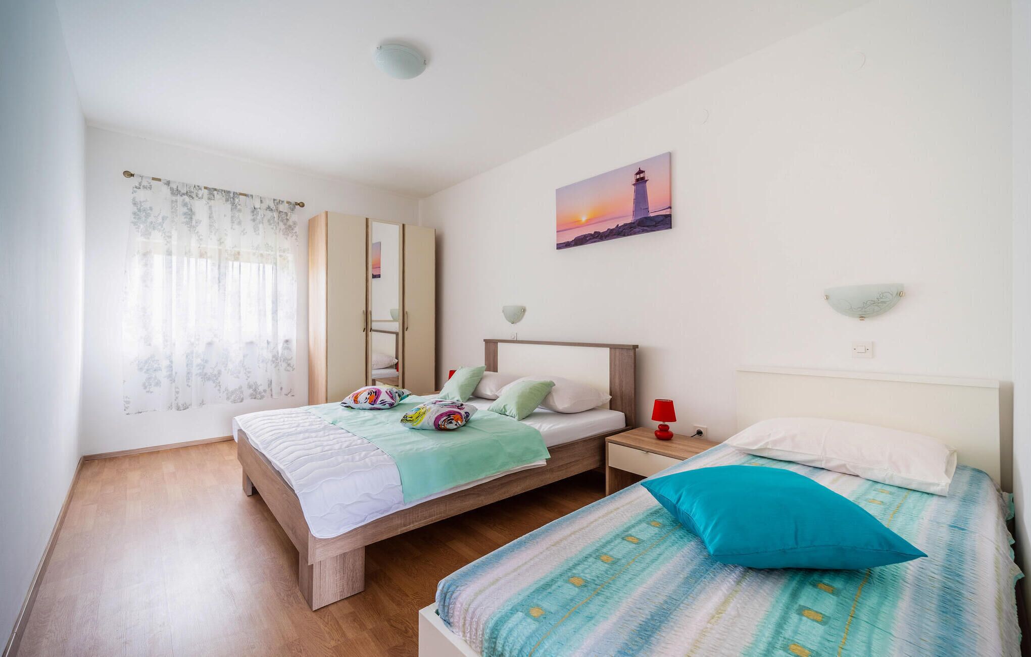 A bedroom with two beds, a wardrobe, a painting of a lighthouse, and a window with curtains.